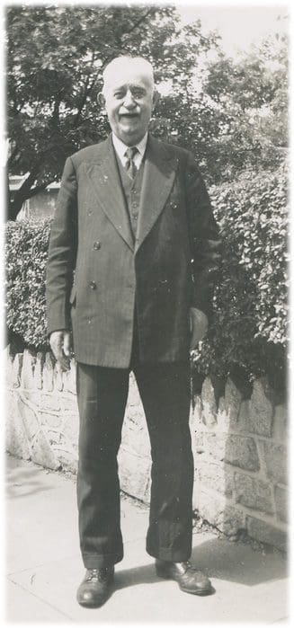 A man in suit and tie standing next to bushes.