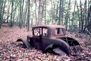 A rusted old car in the woods with leaves on it.