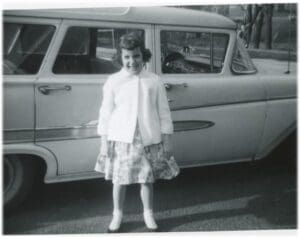 A young girl standing in front of an old car.