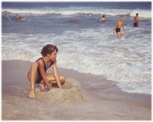 A woman in black bikini sitting on sand near body of water.