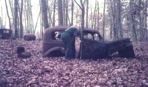 A man in green shirt and blue pants standing next to an old car.